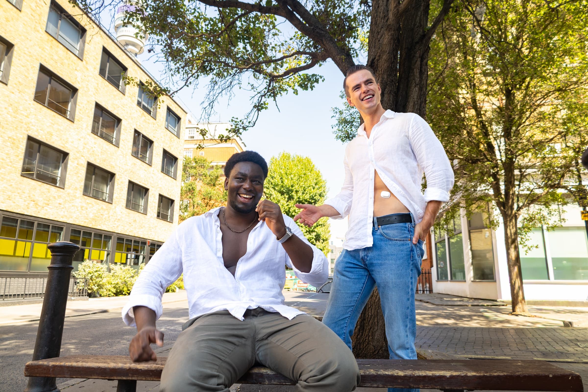 Two models in white shirts laughing on a bench in a city square, one wearing the Suna device